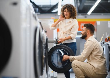 Çamaşır makinesinin özelliklerini inceleyen çift, ev aletleri mağazasında alışveriş yaparken, evleri için satın alma kararlarını birlikte veriyorlar.