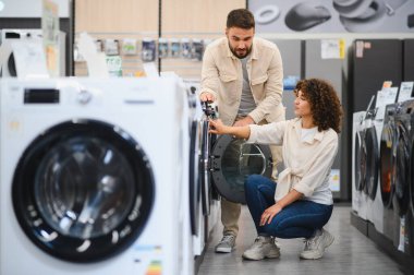 Bir perakende mağazasında ev aletleri seçen çift, çamaşır makineleri ve kurutma makineleri için çeşitli seçenekleri incelerken, özellikleri tartışırken