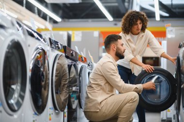 Genç çift, bir mağazada çeşitli çamaşır makinelerini karıştırıyor ve ev çamaşırları için düşünceli kararlar veriyorlar.