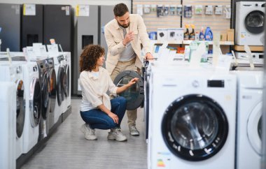Genç çift canlı bir ev aletleri mağazasında yeni bir çamaşır makinesinin seçeneklerini tarıyor ve karşılaştırıyor, özellikleri ve fiyatları tartışıyor