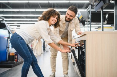 Ev aletleri mağazasında modern fırını inceleyen mutlu çift mutfak malzemeleri için satın alma kararı alıyor.