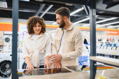Mutlu çift aydınlık bir elektronik mağazasında ev aletleri seçerken bir indüksiyon sobasını işaret ediyor.