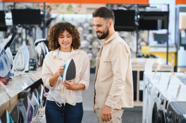 Gülümseyen bir çift elektronik mağazasında yeni bir ütü arıyor. Kadın ütüyü tutarken erkek yararlı tavsiyeler veriyor.