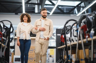 Genç bir çift elektronik mağazasında el ele yürüyorlar. Evleri için yeni bir elektrikli süpürge seçiyorlar.
