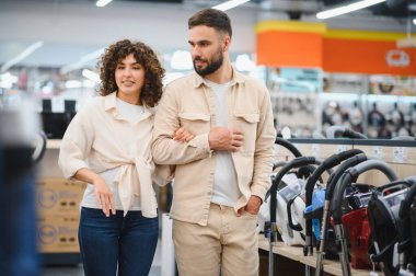 Genç çift ev aletleri mağazasında alışveriş yaparken çeşitli elektrikli süpürgelere göz atıyorlar, seçenekleri tartışıyor ve kararlar alıyorlar.