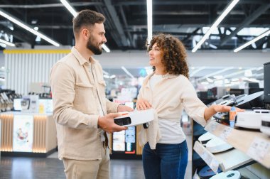 Genç çift yeni robot elektrik süpürgesi seçiyor, büyük bir elektronik mağazasında birlikte alışveriş yapıyorlar.