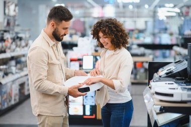Elektrikli süpürgeyi inceleyen mutlu bir çift kalabalık bir elektronik mağazasında ev aletlerini seçerken alışveriş deneyimlerinin tadını çıkarıyor.