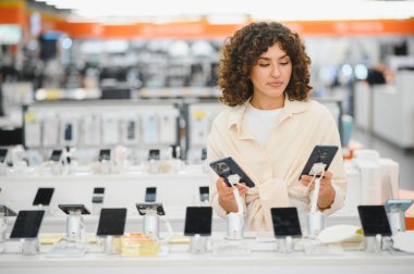 Kadın, tüketici elektroniği mağazasında alışveriş yaparken iki akıllı telefonu karşılaştırıyor ve en iyi cep telefonu anlaşmasını seçiyor.