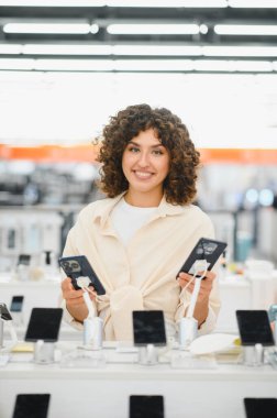 Gülümseyen kadın bir elektronik mağazasında iki akıllı telefonu karşılaştırıyor. Etrafı diğer cep telefonlarıyla çevrili.