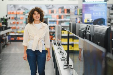 Gülümseyen kadın bir elektronik mağazasında geziniyor, ev eğlencesi için yeni bir televizyon seçerken seçenekleri tarıyor ve karşılaştırıyor.