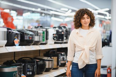 Genç bir kadın, çeşitli modern mutfak teknolojileri ile çevrili, hareketli bir ev aletleri mağazasından yeni bir çoklu pişirici seçiyor.