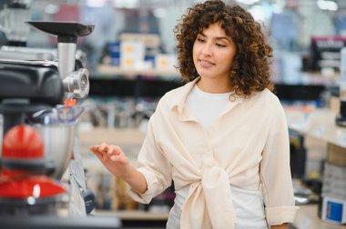 Genç bir kadın bir elektronik mağazasında mutfak aletlerini karıştırıyor, çeşitli ürünlere işaret ediyor ve kendi seçimini yapıyor.