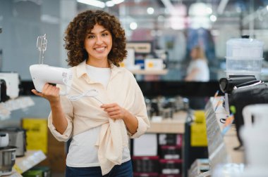 Canlı bir ev aletleri mağazasında mutfak malzemelerini karıştırırken gülümseyen bir kadın alışveriş deneyiminin tadını çıkarıyor.