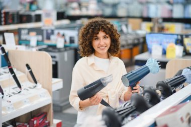Bir elektronik mağazasında ev aletleri alırken iki saç kurutma makinesini karşılaştıran gülümseyen kadın, seçim sürecinin tadını çıkarıyor.