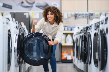 Parlak, modern bir ev aletleri mağazasında çamaşır makinesini seçerken gülümseyen bir kadın memnuniyetini gösteriyor.