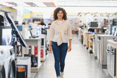 Genç bir kadın elektronik mağazasında yürüyor, ev aletlerini seçiyor, gülümsüyor ve alışveriş deneyiminden zevk alıyor.