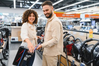 Gülümseyen çift elektronik mağazasında alışveriş yaparken çeşitli elektrikli süpürgelere göz atıyorlar, zamanlarının tadını çıkarıyorlar ve kararlarını veriyorlar.