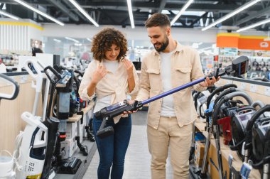 Elektronik mağazasından kablosuz elektrik süpürgesi seçen genç çift, farklı modellerin özelliklerini ve faydalarını tartışıyor.