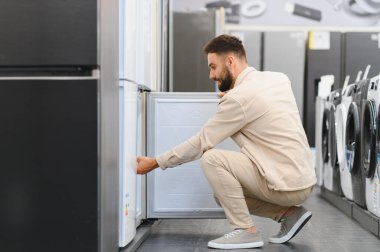 Ev aletleri mağazasında çömelmiş ve buzdolabının kapısını açan sakallı adam, yeni buzdolabı seçiyor.