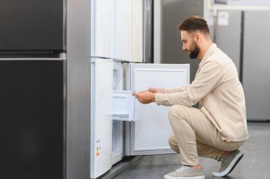 Sakallı adam, elektronik mağazasında yeni ev aletlerini seçerken çömelmiş ve buzdolabının kapısını açıyor.