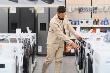 Sakallı adam yeni çamaşır makinesi seçiyor, özelliklerini inceliyor ve modelleri karşılaştırıyor.