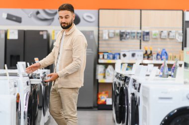 Sakallı adam alışveriş yapmadan önce yeni çamaşır makinesi seçiyor, özelliklerini ve modellerini karşılaştırıyor.