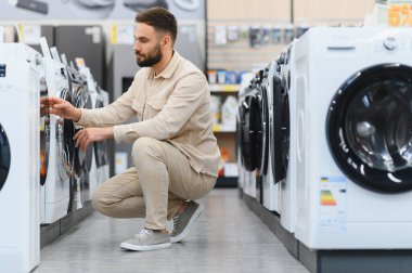 Sakallı bir adam bir ev aletleri mağazasında çömelmiş, dikkatle yeni bir çamaşır makinesi seçiyor.
