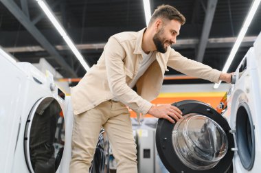 Sakallı adam yeni çamaşır makinesi seçiyor, özelliklerini inceliyor ve satın alma kararı veriyor.