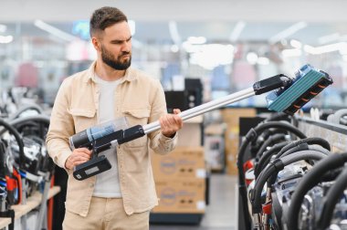 Ev aletleri mağazasında alışveriş yaparken çeşitli kablosuz elektrik süpürgelerini inceleyen ve karşılaştıran sakallı bir adam bir karar vermeye odaklanmış.