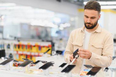 Tüketici elektroniği mağazasından yeni cep telefonu seçen sakallı adam, vitrindeki çeşitli modelleri inceliyor