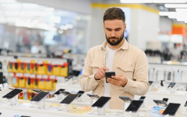Sakallı adam akıllı telefonunu tüketici elektronik mağazasındaki cep telefonu özelliklerini karşılaştırmak için kullanıyor.