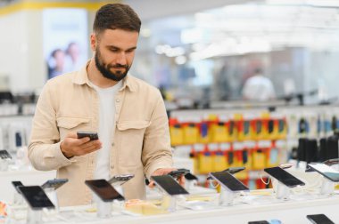 Elektronik mağazasında akıllı telefonu seçen sakallı adam, özelliklerini ve fiyatlarını karşılaştırmak için cep telefonunu kullanıyor.
