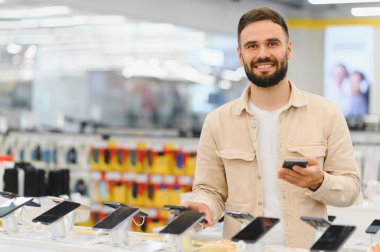 Gülümseyen müşteri yeni bir cep telefonu seçeneklerini karşılaştırırken bir elektronik mağazasında seçimi tarıyor, alışveriş deneyiminin tadını çıkarıyor