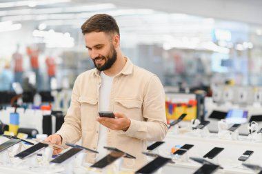 Gülümseyen müşteri yeni akıllı telefonları karşılaştırıyor ve bir elektronik mağazasında telefonundan eleştiriler okuyor.