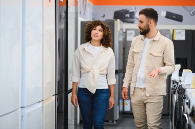 Elektronik mağazasında yürüyen genç bir çift yeni buzdolabı arıyor, farklı modellerin özelliklerini ve faydalarını tartışıyorlar.