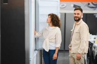 Eşyalar mağazasında çeşitli buzdolabını karıştıran çift seçeneklerini karşılaştırıyor ve mutfak güncellemelerinin özelliklerini tartışıyor.