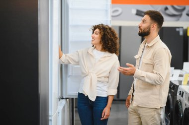 Çift bir elektronik mağazasında buzdolaplarını inceliyor, seçenekleri ve özellikleri tartışıyorlar, aynı zamanda evleri için bir satın alma kararı veriyorlar.