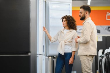 Genç çift eşyalar mağazasında yeni buzdolabı seçiyor, kadın buzdolabının kapısını açıyor ve partneriyle özellikleri tartışıyor.