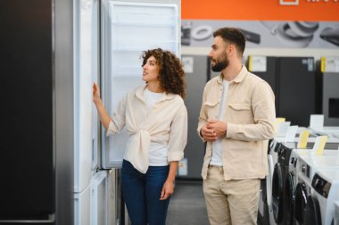 Çift, ev aletleri seçeneklerini göz önünde bulundurarak elektronik mağazasında bir buzdolabını inceliyor. Odaklanmış ve karar vermeye odaklanmış görünüyorlar.