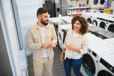 Mutlu çift seçenekleri tartışıyor ve modern ev aletleri mağazasında alışveriş yaparken buzdolabının özelliklerini karşılaştırıyor