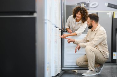 Bir çift yeni bir buzdolabı seçiyor, seçenekleri tartışıyor ve modern mutfak güncellemeleri için özellikleri karşılaştırıyor.