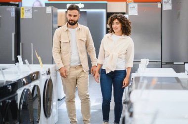 Genç çift el ele tutuşup, modern, parlak bir ev aletleri mağazasında çamaşır makinesini seçiyorlar. Birlikte alışveriş deneyimlerinin tadını çıkarıyorlar.