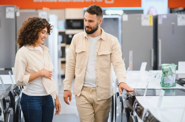 Genç çift çamaşır makinelerini karıştırıyor, seçenekleri tartışıyor ve elektronik mağazasından ev aletleri alırken seçimler yapıyor.