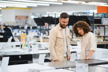 Çift, satın alma seçeneklerini göz önünde bulundurarak bir elektronik mağazasında ev aletlerini incelerken görülüyor. Odaklanmış ve meşgul görünüyorlar.