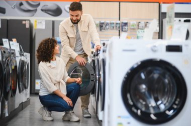 Genç bir çift ev aletleri mağazasında yeni bir çamaşır makinesi seçiyor, özelliklerini inceliyor ve seçeneklerini tartışıyor.