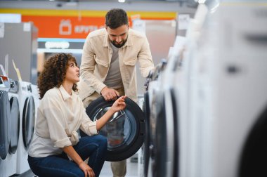 Geniş bir ev aletleri mağazasında yeni bir çamaşır makinesi seçen genç çift, özellikleri tartışıyor ve satın alma kararı alıyorlar.