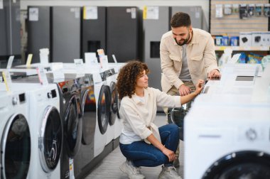 Çift bir elektronik mağazasında çeşitli ev aletlerini inceler, seçenekleri tartışır ve satın almaları hakkında kararlar verir.