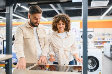 Ev aletleri mağazasında mutfak aletlerini seçen mutlu çift, modern bir indüksiyon sobasına bakıyor.