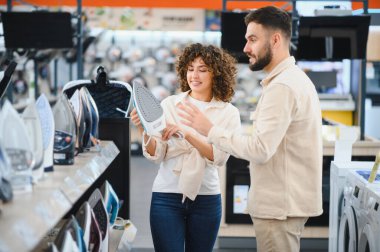 Genç bir çift elektronik mağazasında yeni bir ütü seçiyor, kadın erkeğe modern bir alet gösteriyor ve satın almayı tartışıyor.