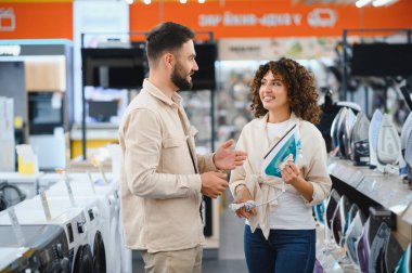 Çift bir elektronik mağazasında alışveriş yapıyor, çeşitli ev aletlerini inceliyor ve satın alma seçeneklerini tartışıyor.